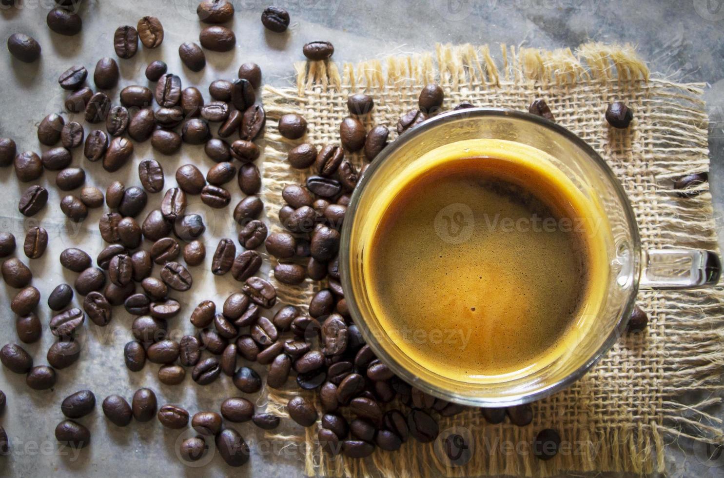 xícara de café americano preto quente em uma mesa de concreto rústica de pano de saco com uma pilha de grãos de café arábica marrom torrado. atualizar, conceito de bebida matinal foto