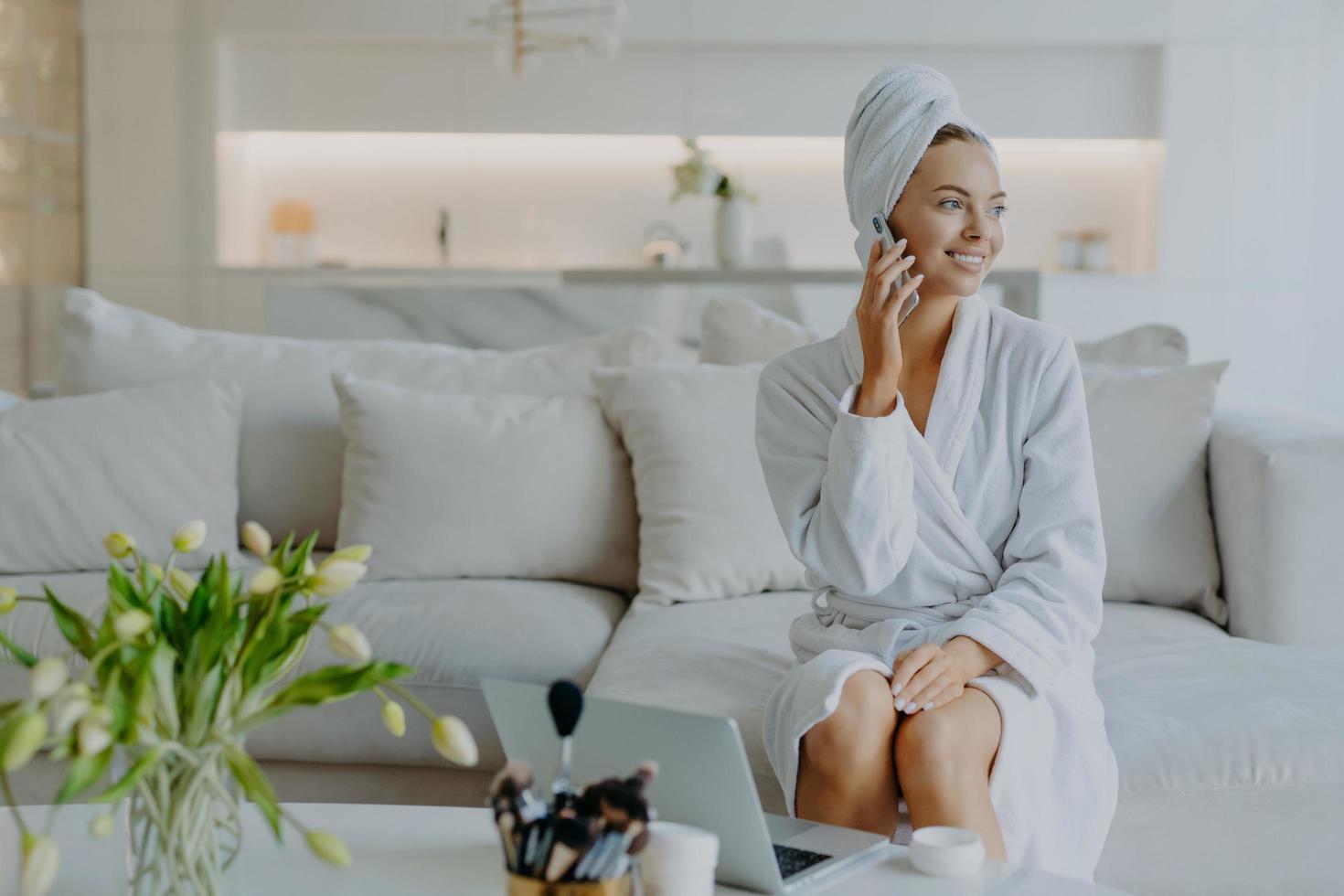 mulher bonita alegre olha de lado com sorriso cheio de dentes tem conversa telefônica vestida de roupão de banho e toalha enrolada na cabeça cercada de dispositivos modernos usa produtos cosméticos para se preocupar consigo mesma foto