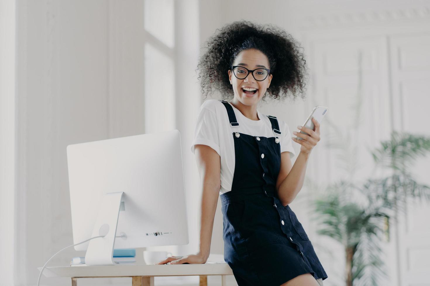 feliz financista feminina despreocupada usa telefone celular, feliz por receber salário a tempo, posa perto de mesa de madeira, usa computador para trabalhar, veste camiseta branca e sarafan, mensagem de texto, sorri agradavelmente foto