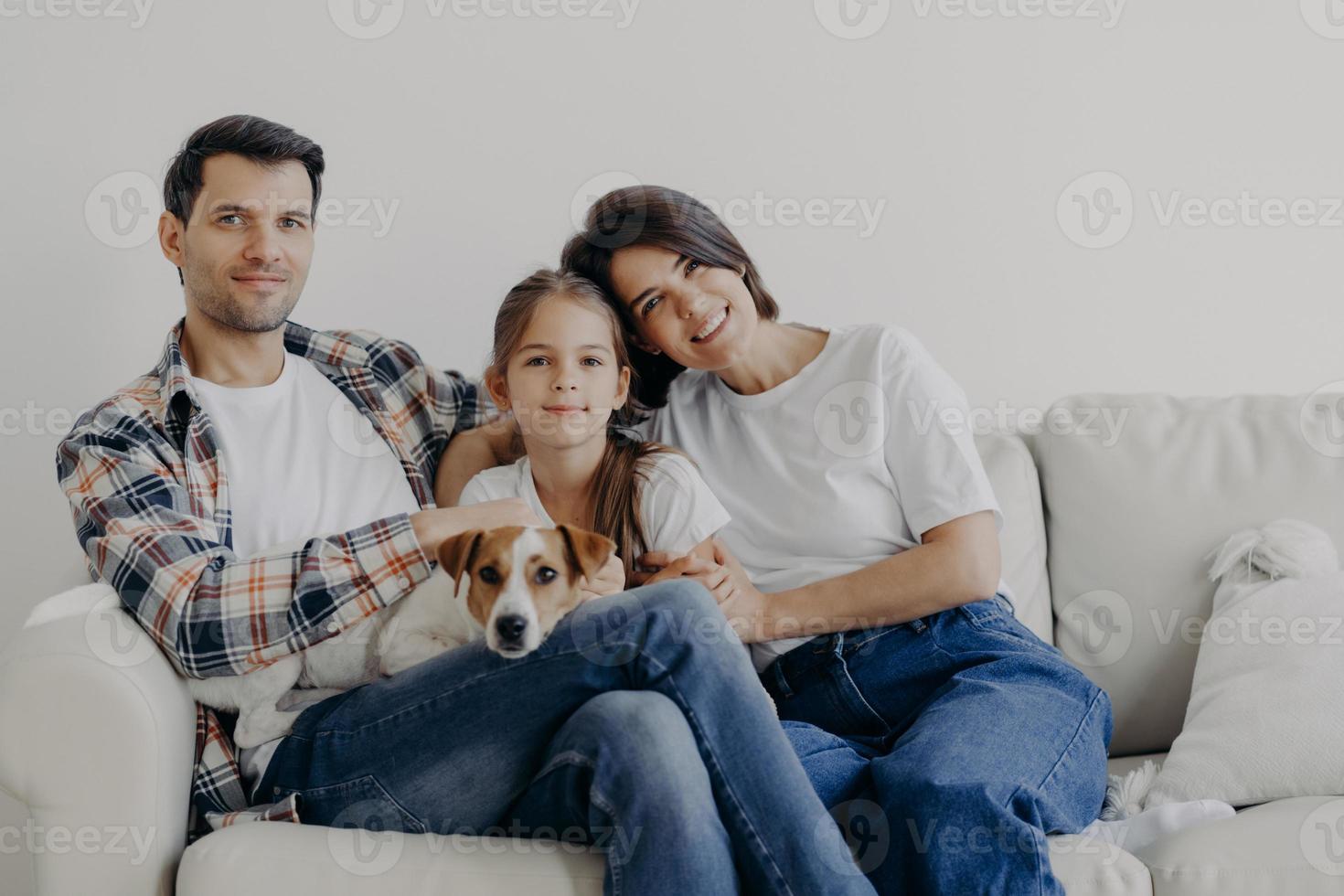 retrato de família afetuosa abraçam-se e sentam-se juntos no sofá na sala de estar, mudam de casa, têm expressões felizes. pai, mãe, filha e cachorro posam para fazer retrato, passam bons momentos foto