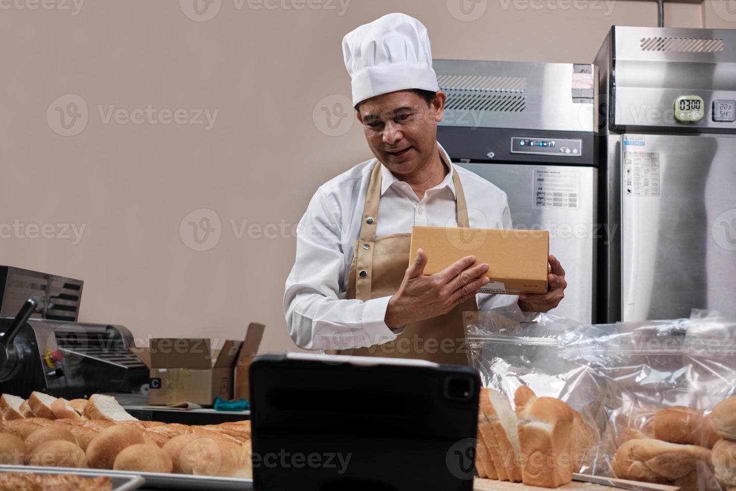 chef masculino asiático sênior de avental, blogueiro de pequenas empresas transmitindo ao vivo via tecnologia de aplicativo móvel, promoção on-line de vendas de padaria, pão fresco, comida de pastelaria e culinária comercial na cozinha. foto