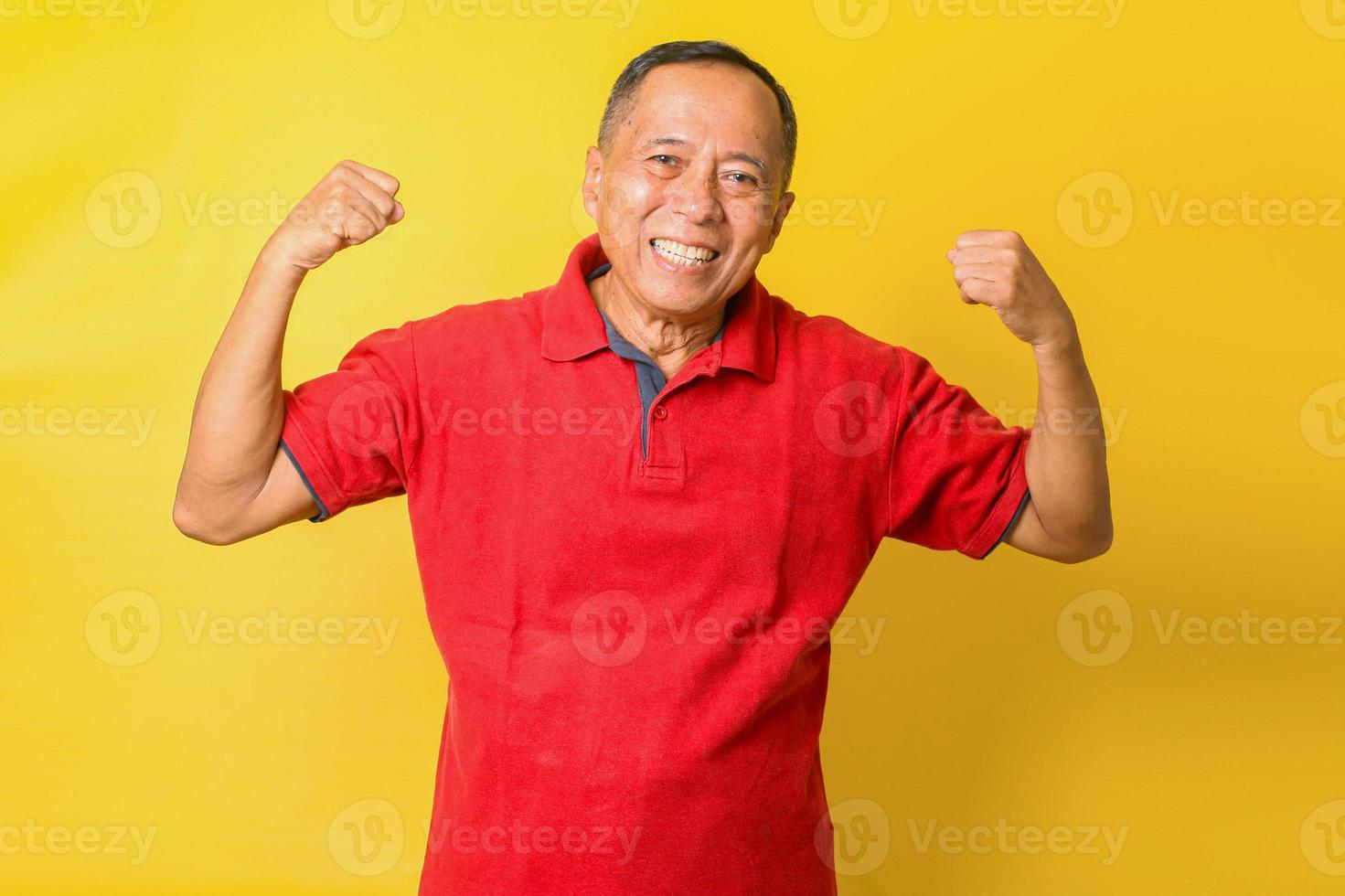 vovô aposentado asiático levanta duas mãos mostra que os músculos usam camiseta vermelha isolado fundo de cor amarela foto