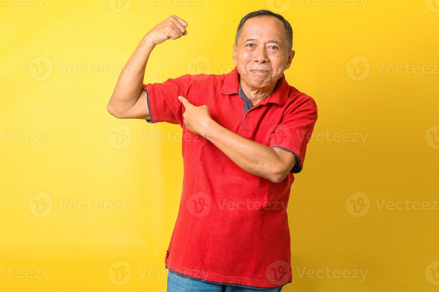 pensionista asiático velho levantar o dedo direto do braço demonstrar bíceps usar camiseta vermelha isolado fundo de cor amarela foto
