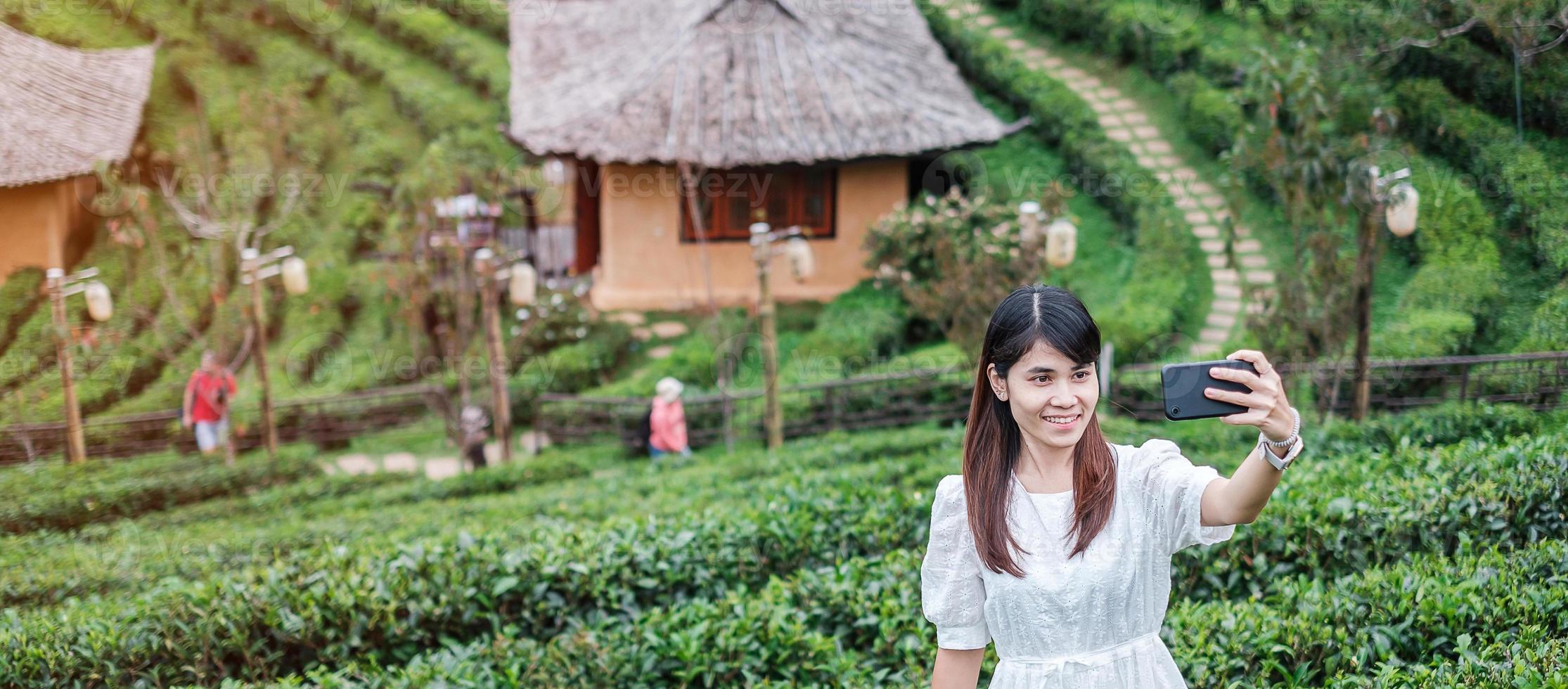 mulher de turista feliz no vestido branco tirando foto pelo smartphone móvel no lindo chá garden.traveler visitando na vila tailandesa de ban rak, mae hong son, tailândia. conceito de viagens, férias e férias