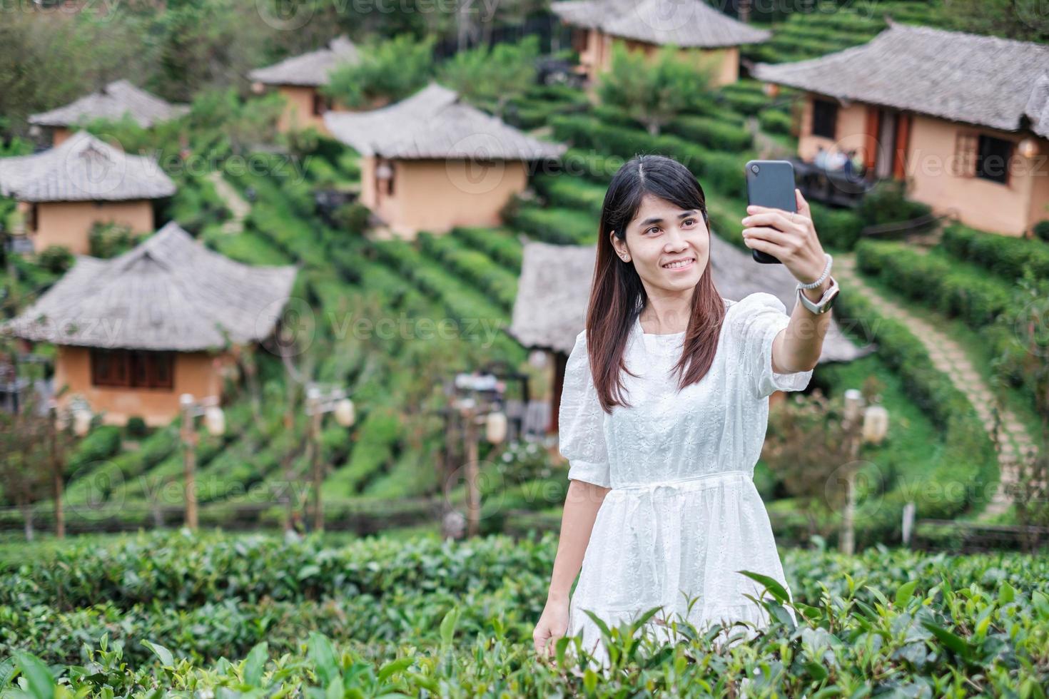 mulher de turista feliz no vestido branco tirando foto pelo smartphone móvel no lindo chá garden.traveler visitando na vila tailandesa de ban rak, mae hong son, tailândia. conceito de viagens, férias e férias