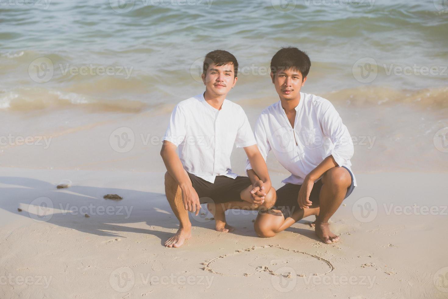 jovem casal asiático gay sorrindo romântico desenho coração forma juntos na areia em férias, homossexual feliz e divertido com amor sentado na areia na praia no verão de viagens, conceito legal lgbt. foto