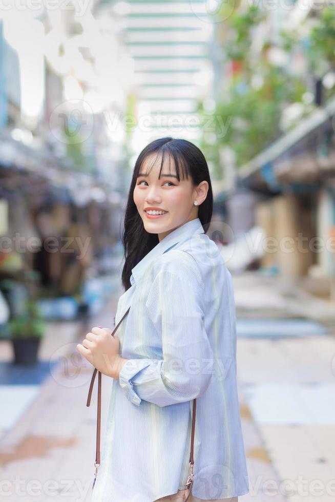 confiante jovem mulher asiática que veste uma camisa branca azul e bolsa sorri alegremente e olha para a câmera enquanto viaja para o trabalho pela cidade velha. foto