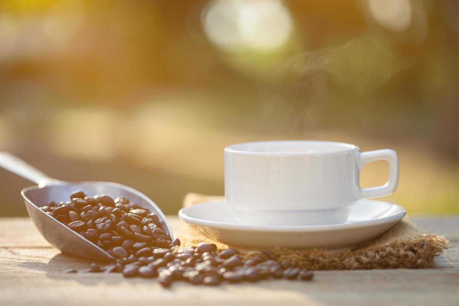 xícara de café e grãos de café na mesa de madeira ao ar livre na luz do sol da manhã e bokeh de fundo foto
