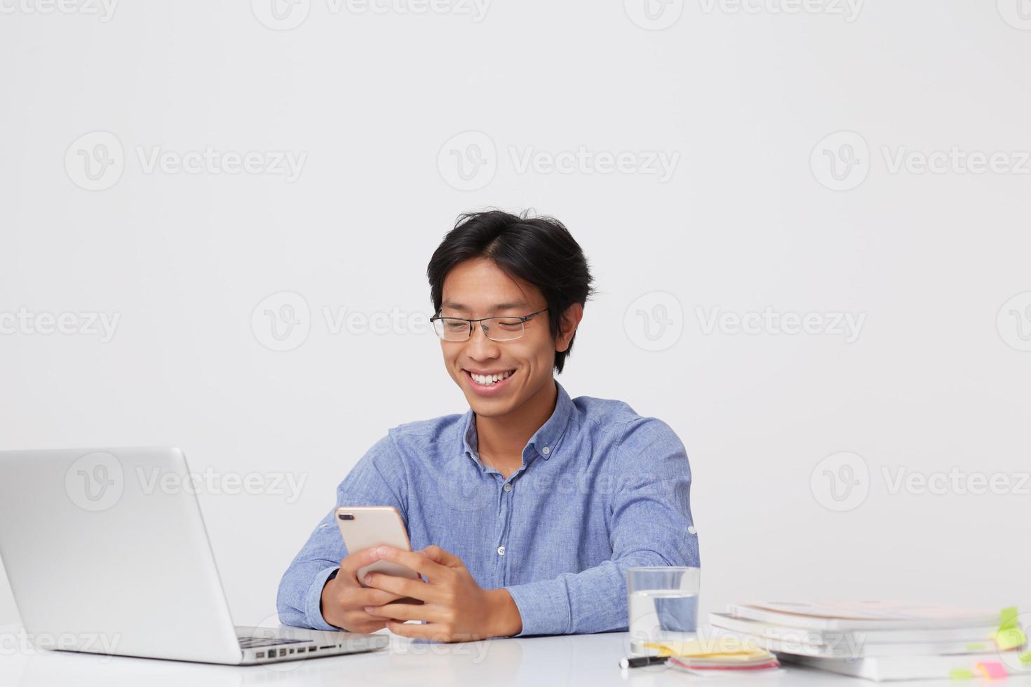 sorrindo bonito jovem empresário asiático de óculos e camisa azul usando telefone celular e laptop sentado à mesa sobre fundo branco foto