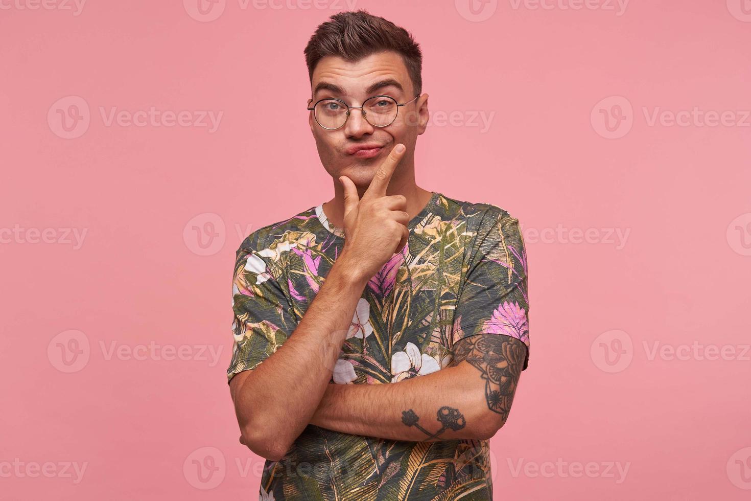 indiferente bonito jovem macho de cabelos escuros dobrando os lábios com sobrancelhas levantadas, segurando o queixo com rosto apático, posando sobre fundo rosa foto