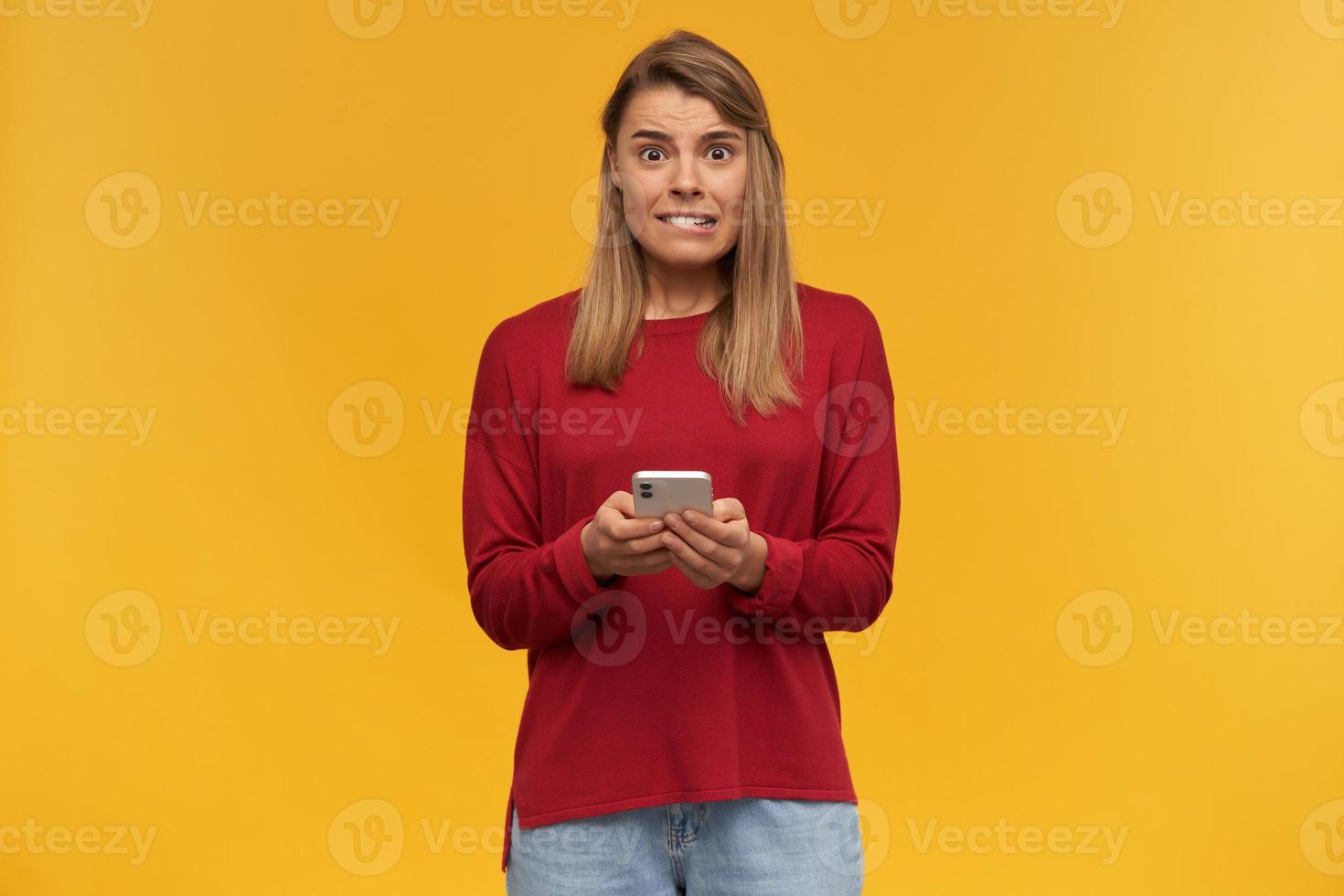 jovem loira parece insegura como se estivesse em apuros ou em uma situação embaraçosa, mantém o celular nas mãos, olhando a câmera com medo, morde um pouco o lábio, em um fundo amarelo do estúdio foto