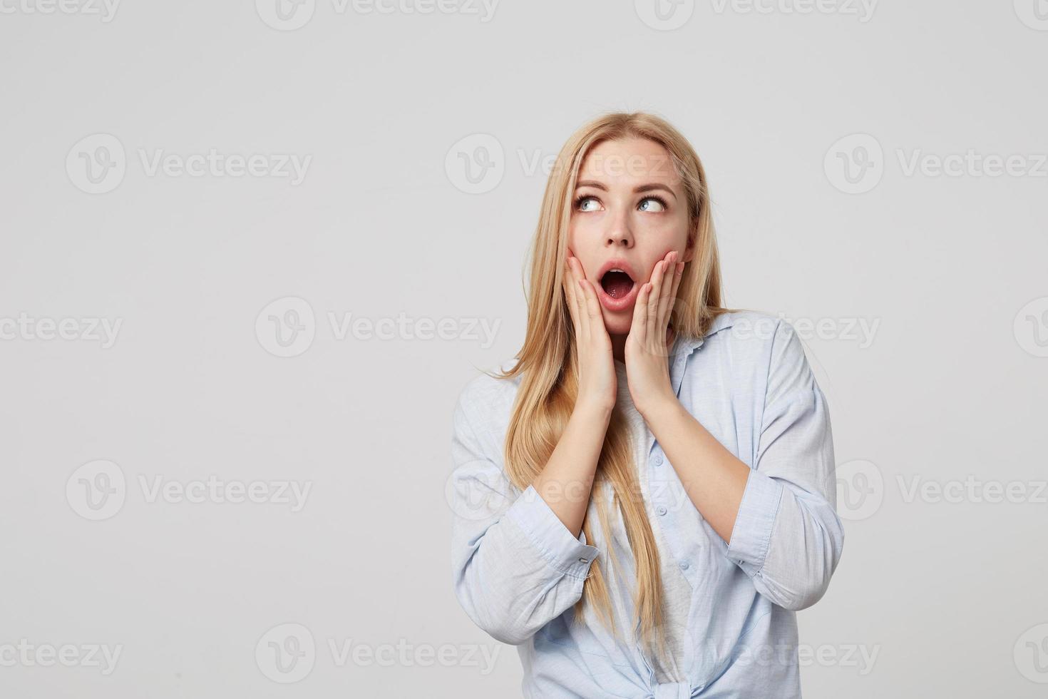 retrato de uma linda loira surpresa com as palmas das mãos nas bochechas, olhando de lado espantado com a boca aberta, de pé sobre fundo branco foto