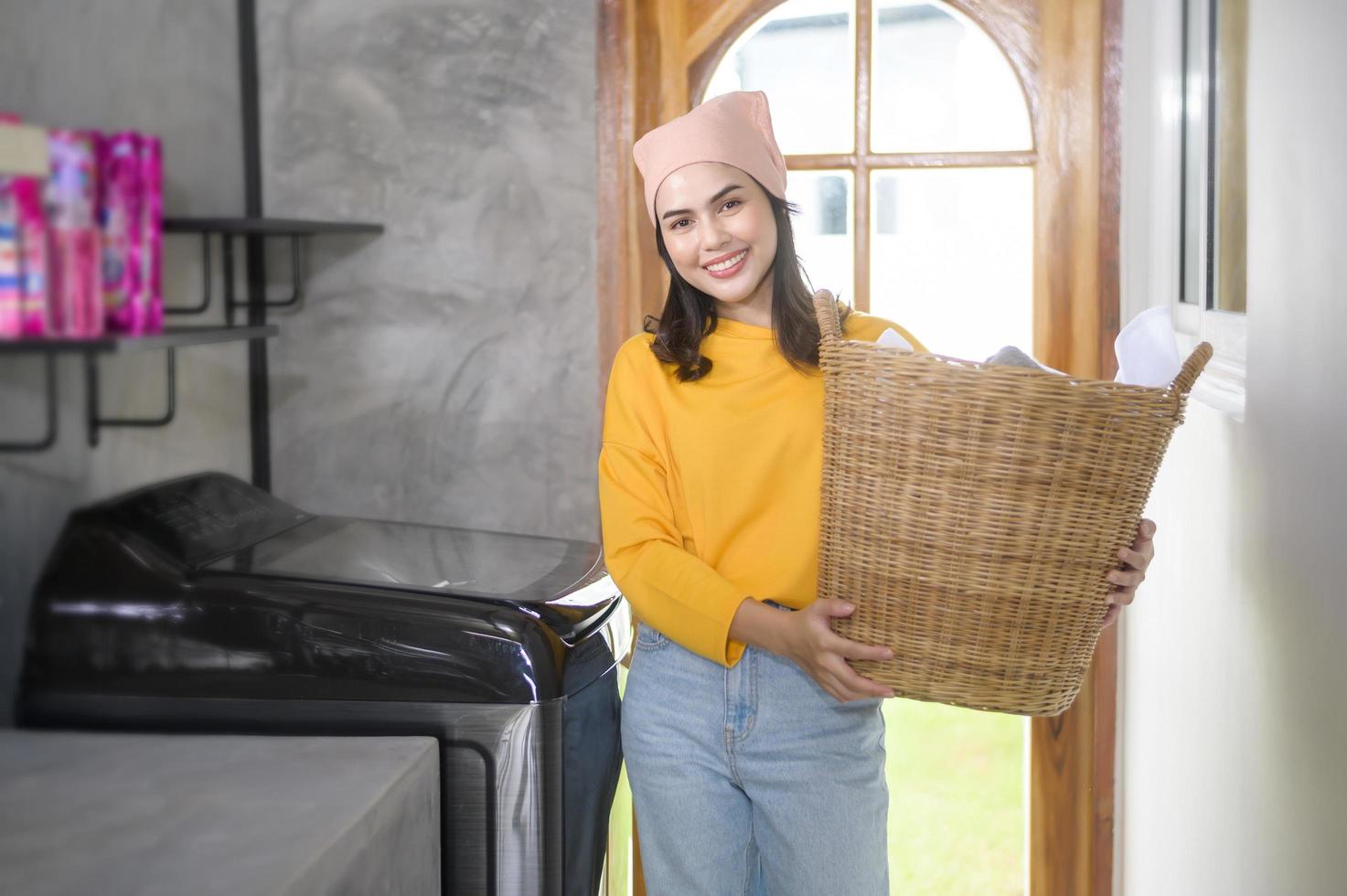 jovem mulher feliz vestindo camisa amarela segurando uma cesta cheia de roupas em casa, conceito de lavanderia foto