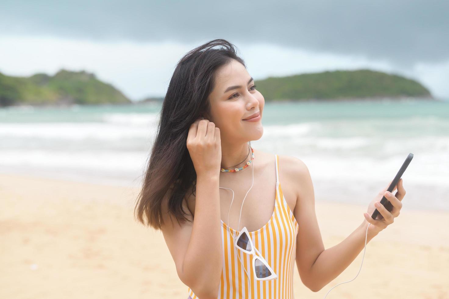 jovem mulher bonita de biquíni ouvindo música e usando smartphone na praia, verão, férias, feriados, conceito de estilos de vida. foto