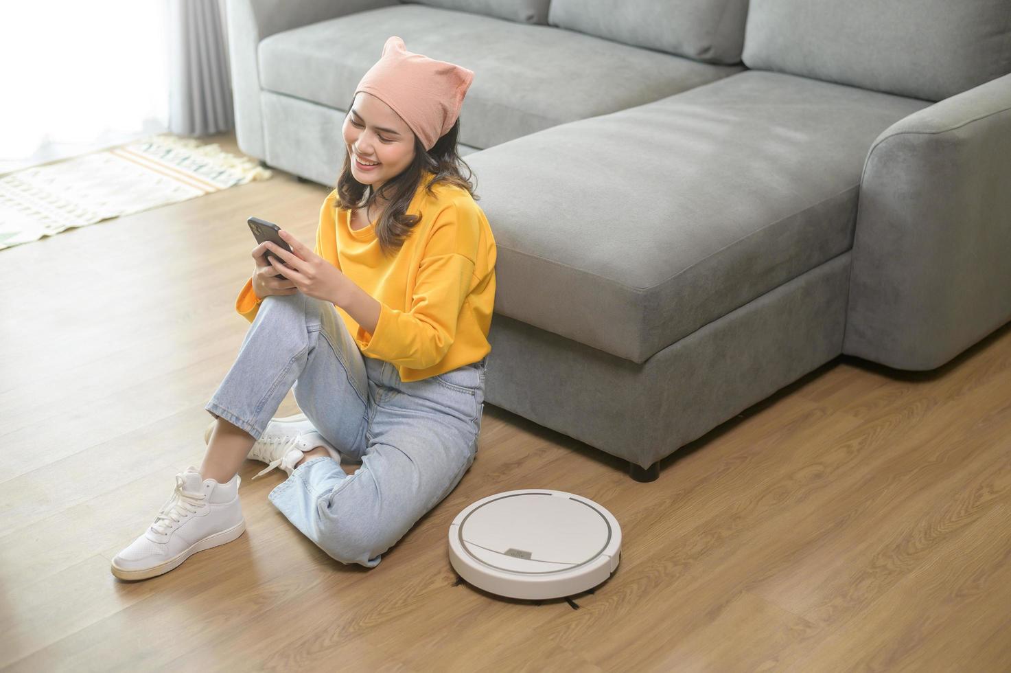 jovem mulher feliz relaxando e usando smartphone na sala de estar enquanto aspirador de pó robótico trabalhando foto