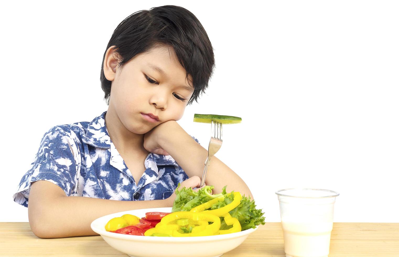 menino adorável asiático mostrando expressão chata com legumes coloridos frescos e copo de leite isolado sobre fundo branco foto