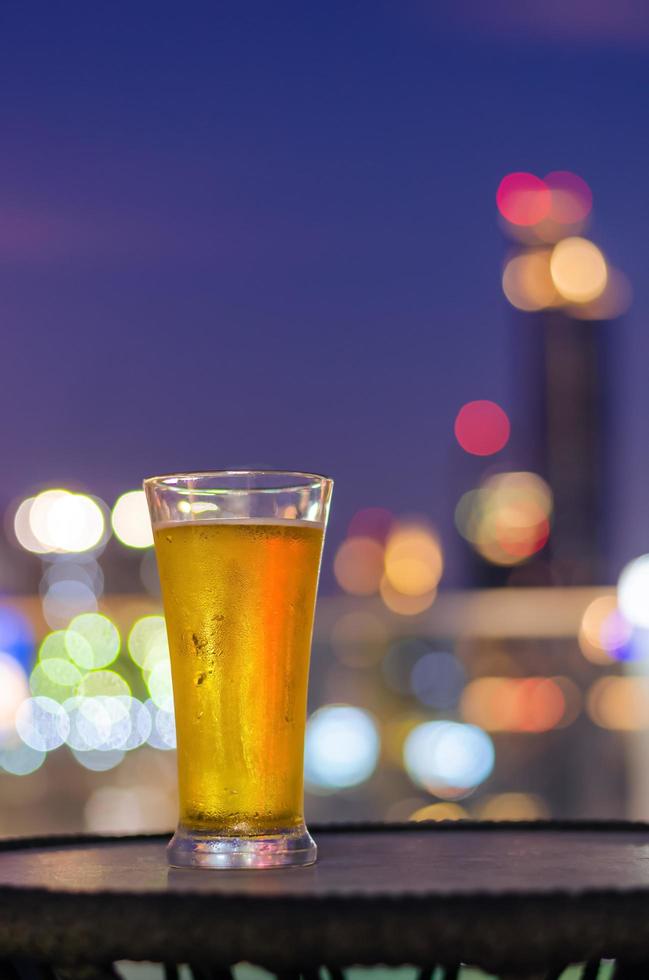 um copo de cerveja coloca na mesa no bar da cobertura com luz de fundo colorido bokeh da cidade. foto