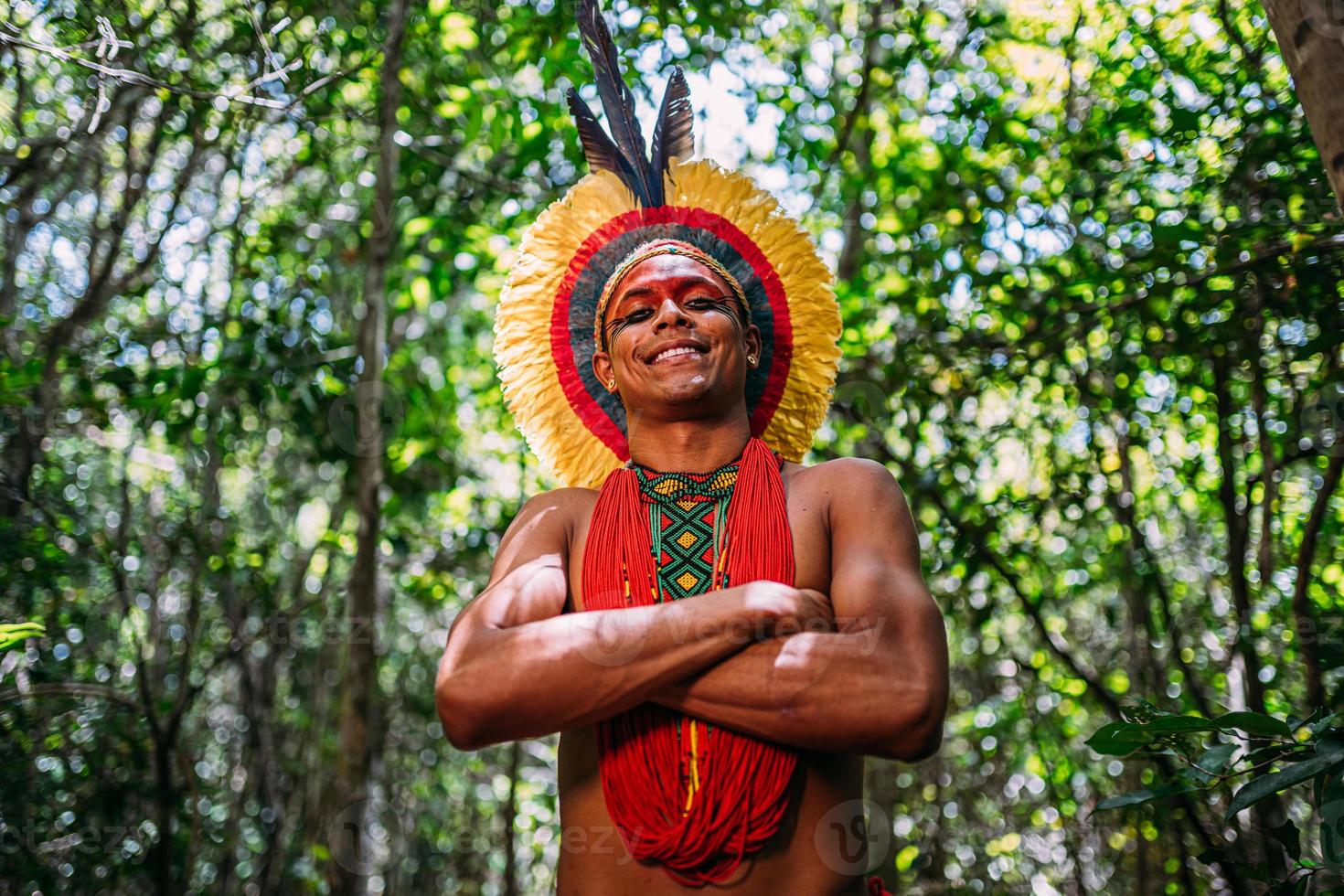 índio da tribo pataxo, com cocar de penas. jovem índio brasileiro olhando para a câmera, sorrindo e braços cruzados foto