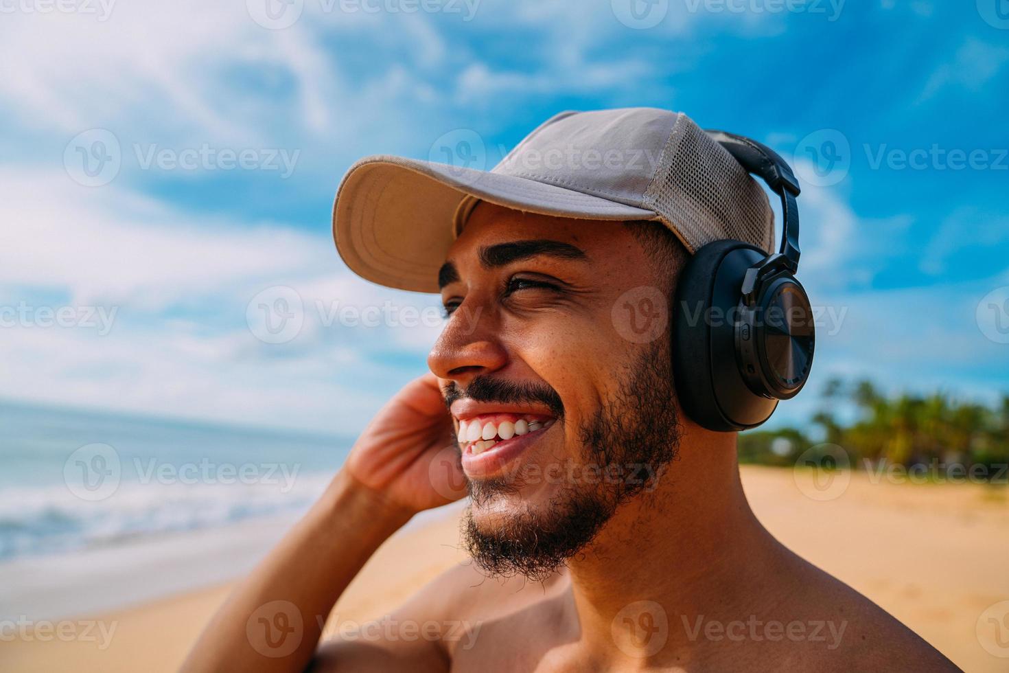 férias de verão, tecnologia e conceito de internet. homem latino-americano olhando para a esquerda, ouvindo música com fone de ouvido e tomando sol na praia foto