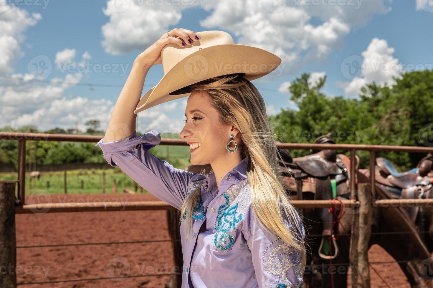 linda vaqueira loira com chapéu de pé perto do fundo do rancho de cavalos foto