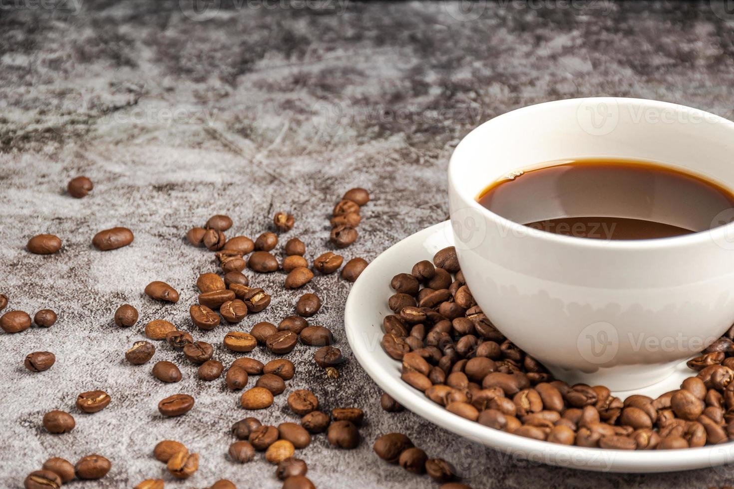 um close-up de café quente em uma xícara branca é colocado em uma mesa de piso de cimento, muitos grãos de café torrados estão no pires da xícara de café e, ao redor, fumaça e aroma flutuam da xícara. fundo desfocado foto