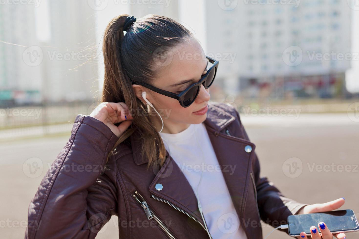 mulher atraente andando na luz do sol em óculos de sol e jaqueta de couro foto