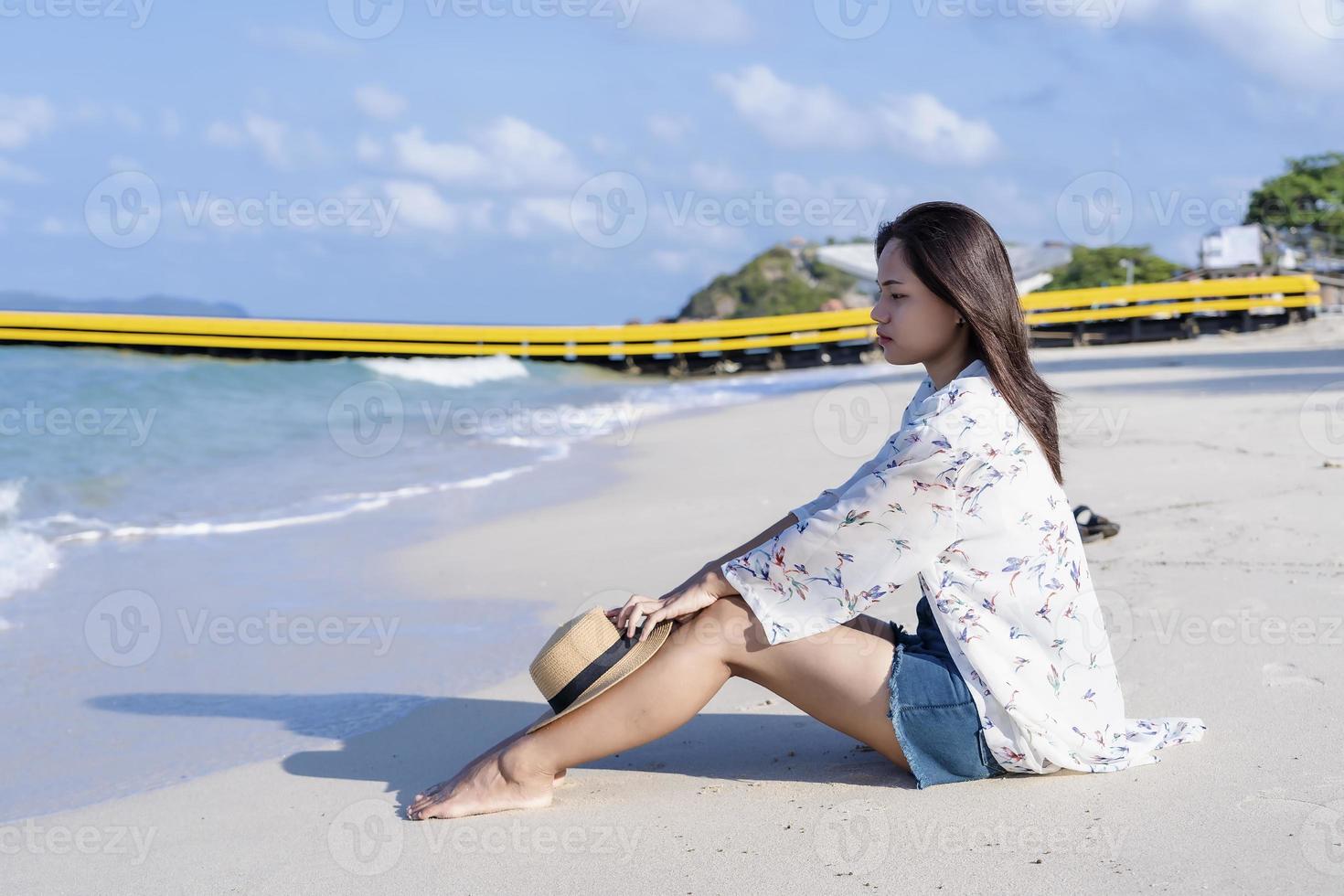 mulher preocupada sentada na praia com um chapéu de palha na praia tropical de joelhos à beira-mar no dia ensolarado. garota solitária sentada sozinha à beira-mar, relaxando e pensando. fotografia de conceito de emoção humana foto