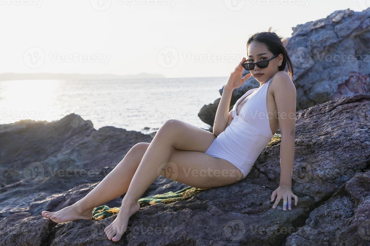 retrato de mulher bonita na moda em biquíni branco posando na praia de pedra. jovem mulher asiática vestida de maiô deitado em uma pedra em um dia de verão com fundos por do sol em koh larn em pattaya foto