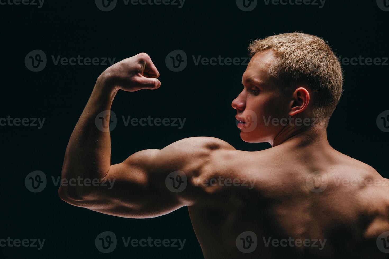 homem jovem musculoso mostrando seus bíceps isolados no fundo preto foto