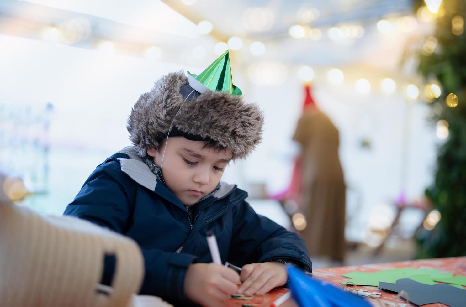 menino bonito com chapéu de festa usando caneta rosa desenhando ou escrevendo na cor do papel com fundo claro embaçado, criança se divertindo na festa de aniversário, criança fazendo atividade no feriado de natal ou ano novo foto