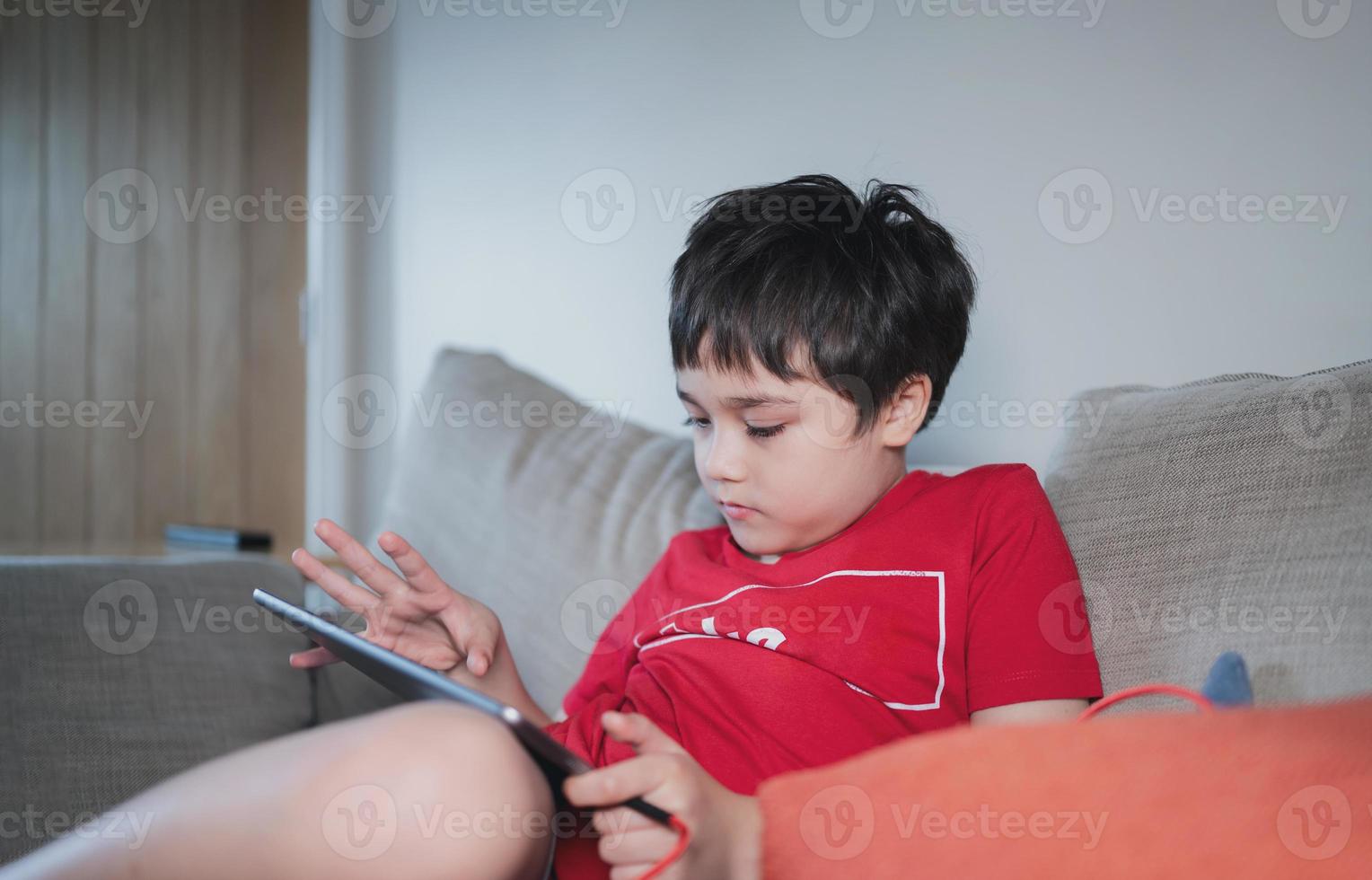 criança sentada no sofá assistindo desenho animado ou jogando no tablet, criança usando almofada digital aprendendo lição on-line na internet, educação em casa, conceito de educação on-line de ensino a distância foto