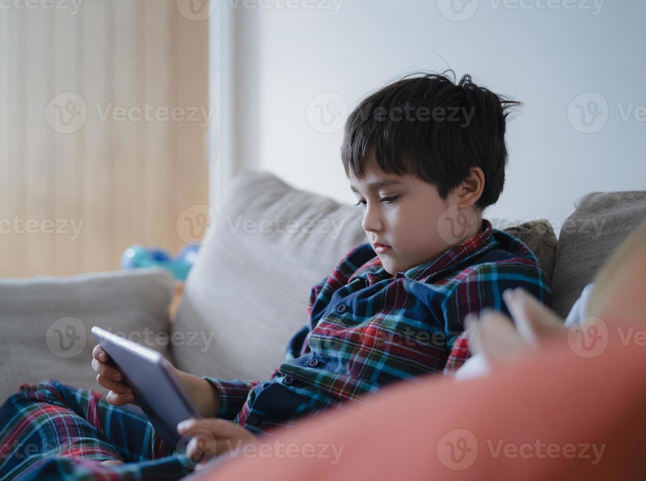 garoto sentado no sofá assistindo desenho animado, menino jogando jogo online no tablet com amigos de férias, menino criança vestindo pijama relaxando em casa pela manhã foto