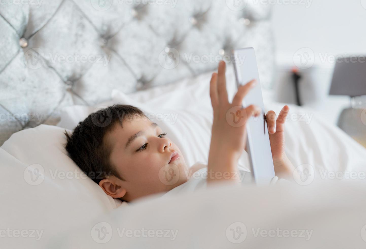 criança feliz, deitada na cama segurando o tablet assistindo desenhos animados e conversando com amigos no teclado digital, menino bonitinho jogando jogos online na internet, criança relaxando de manhã antes de ir para a escola foto