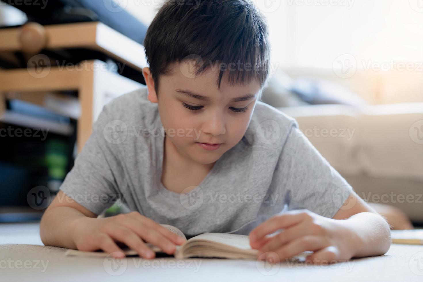 garoto de escola de retrato lendo um livro para lição de casa, menino deitado no tapete gosta de ler a história na sala de estar, criança relaxando em casa no fim de semana. educação em casa ou conceito de educação foto