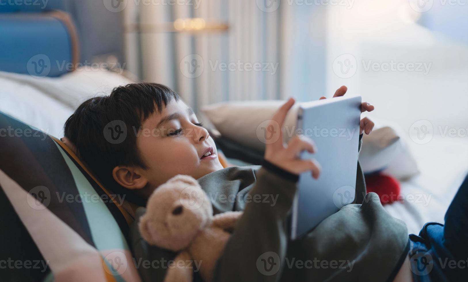 criança feliz, deitada na cama segurando o tablet assistindo desenhos animados e conversando com amigos no teclado digital, menino bonitinho jogando online na internet, criança relaxando em casa com a família foto