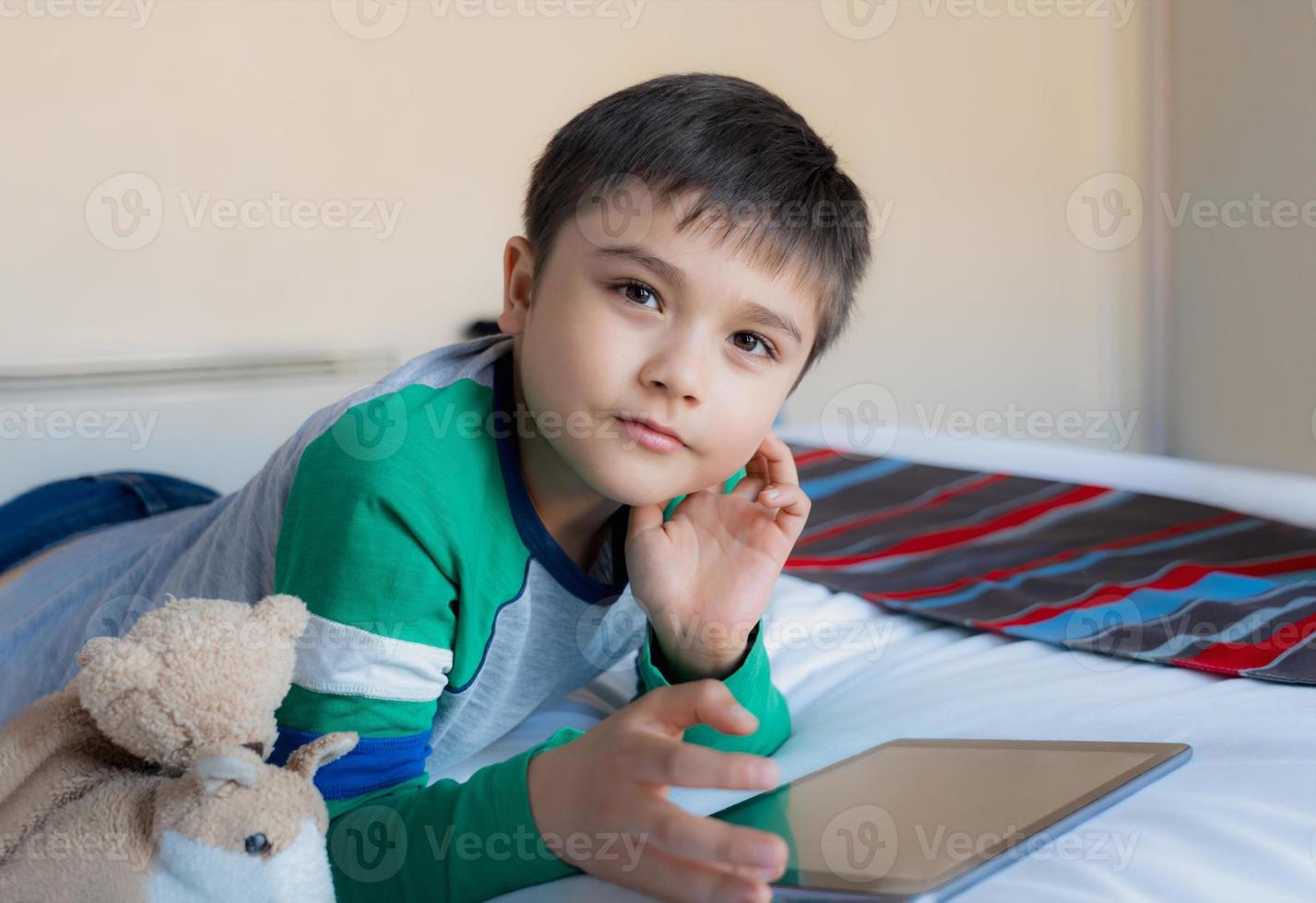 garoto de retrato jogando jogos on-line na internet com amigos no quarto de cama, menino usando tablet digitando ou conversando à noite, infância deitada na cama relaxando em casa no fim de semana. foto