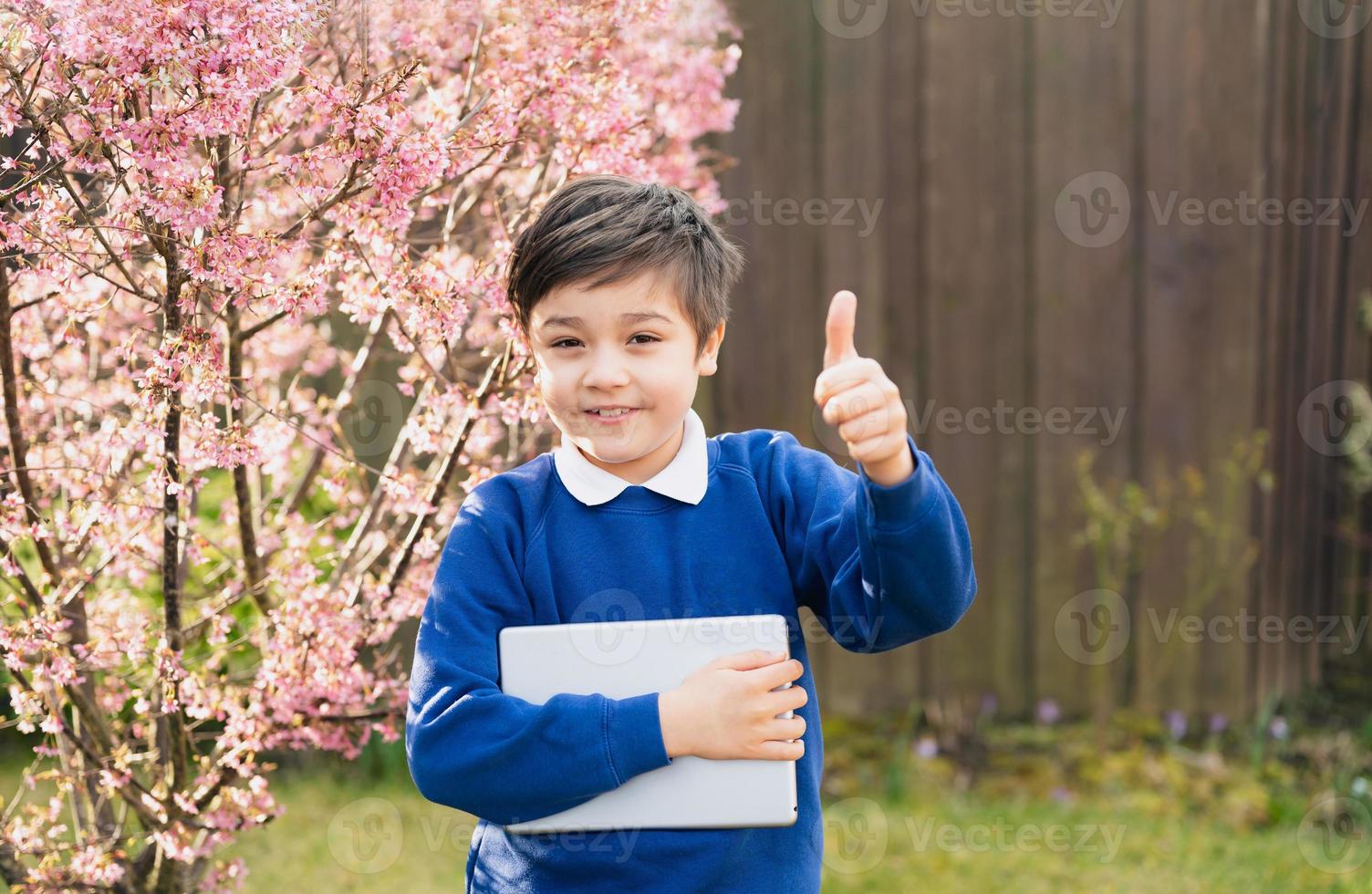 retrato menino feliz mostrando os polegares para cima olhando para a câmera com rosto sorridente, garoto da escola segurando o tablet pc do lado de fora esperando o ônibus escolar na primavera de dia ensolarado foto