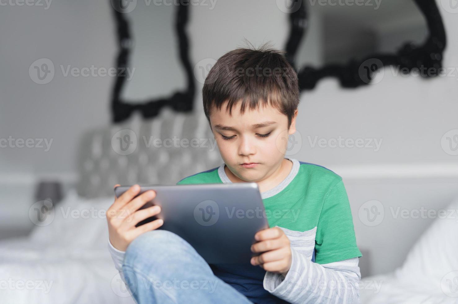 menino usando tablet jogando jogo na internet com um amigo, criança que estuda em casa fazendo lição de casa on-line pelo teclado digital em casa, criança sentada na cama relaxando, assistindo desenhos animados ou conversando online com um amigo foto
