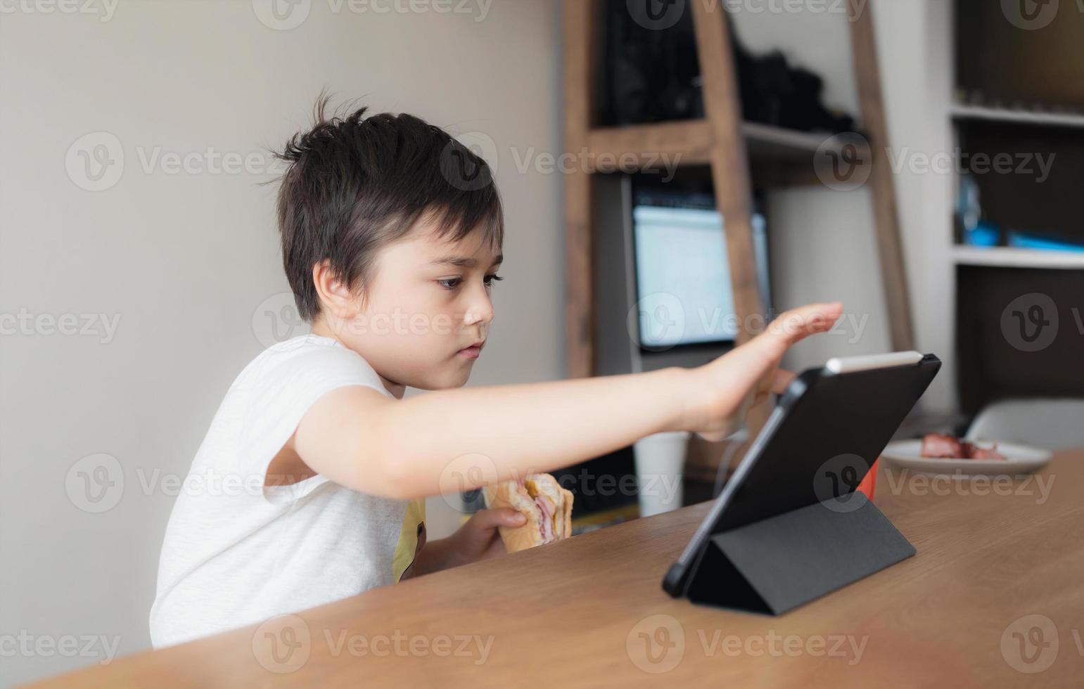 criança jogando jogo online com amigos no tablet, retrato cinematográfico jovem menino chamada de vídeo com amigos enquanto toma café da manhã, criança assistindo desenhos animados na internet, criança da escola fazendo lição de casa no teclado digital foto