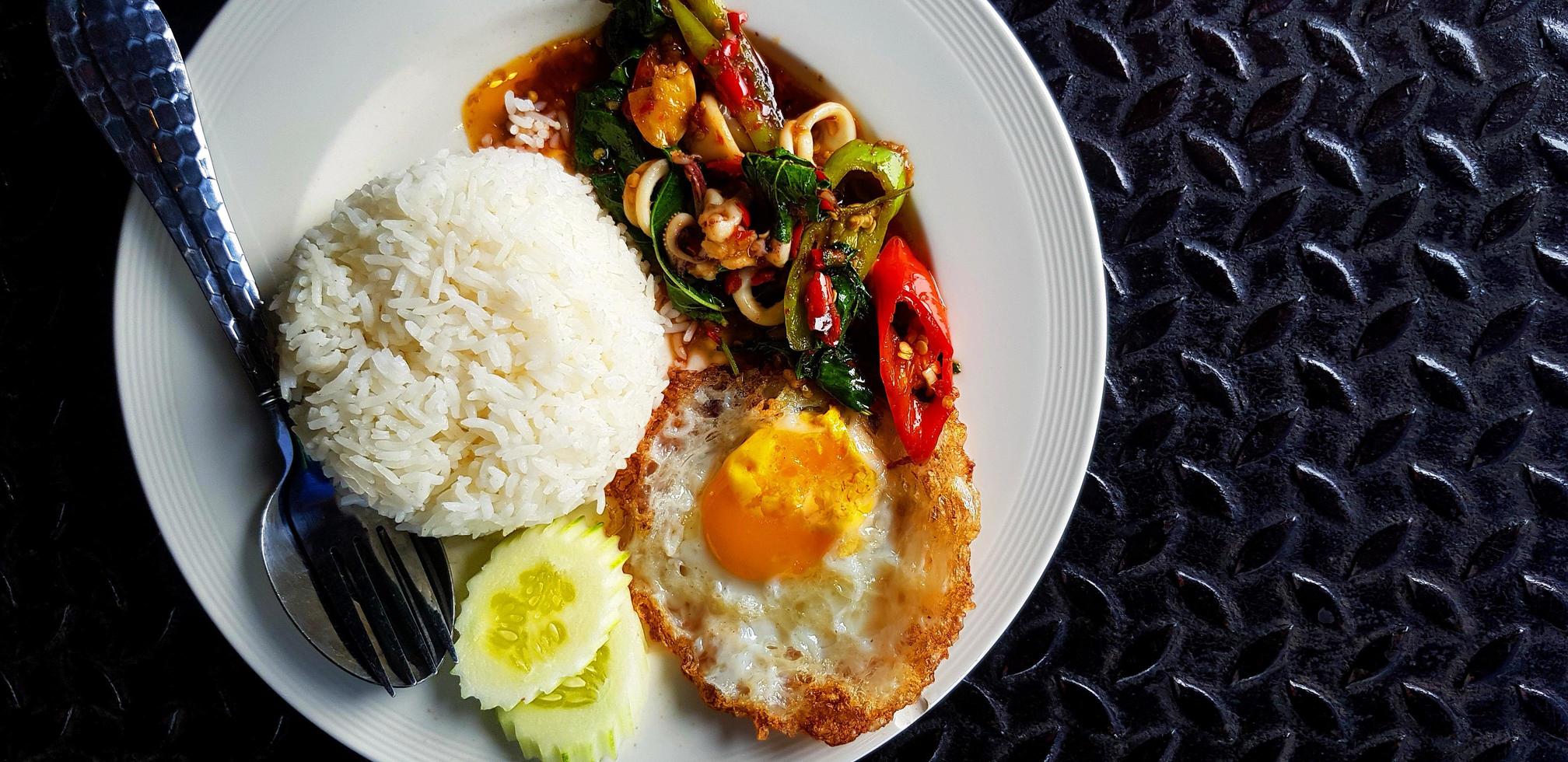 vista superior do manjericão frito com lula, arroz, pepino fatiado e ovo frito em estilo tailandês na chapa branca com espaço de cópia. postura plana de comida famosa e picante em fundo preto de aço inoxidável. foto