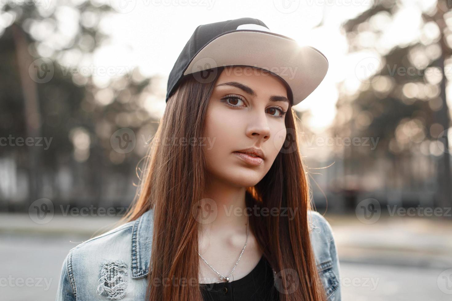 retrato de close-up de uma linda garota hipster em um boné de beisebol preto e jaqueta jeans azul rasgada ao pôr do sol foto