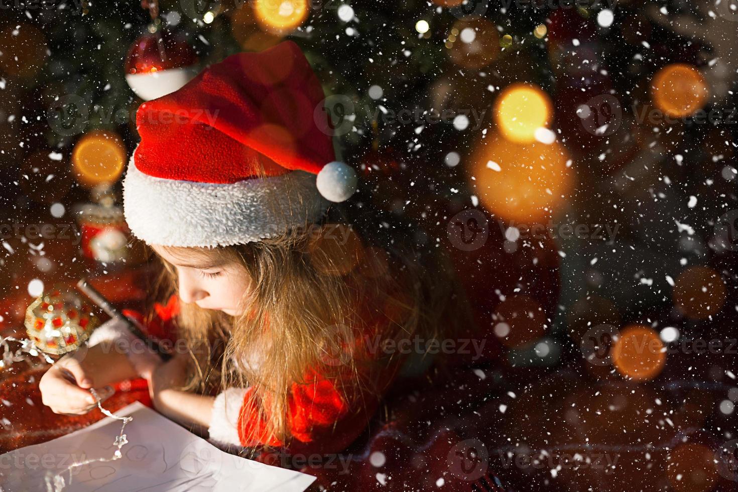 menina com um chapéu de Papai Noel e vestido vermelho debaixo da árvore de natal está sonhando, esperando o feriado, deitado sobre um cobertor xadrez. uma carta em um pedaço de papel, presentes. ano novo, natal. luzes de desfocagem foto