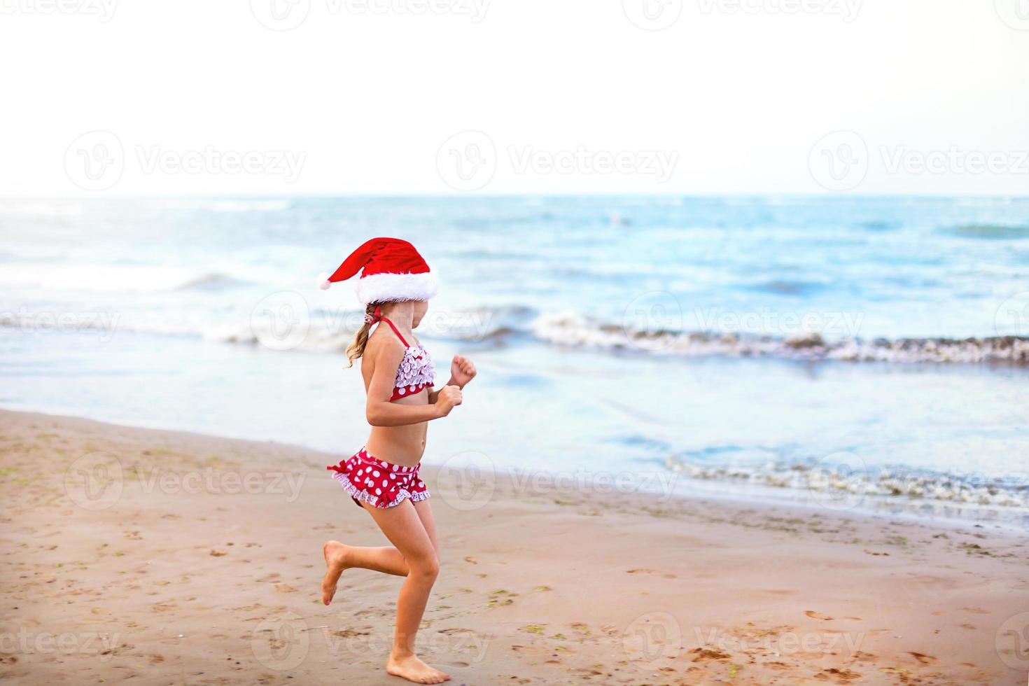 Menina de 5 anos de maiô vermelho e chapéu de papai noel na praia está feliz, grita, pula e acena as mãos com felicidade. natal e ano novo passeios de inverno para países quentes. turismo para férias nos trópicos foto