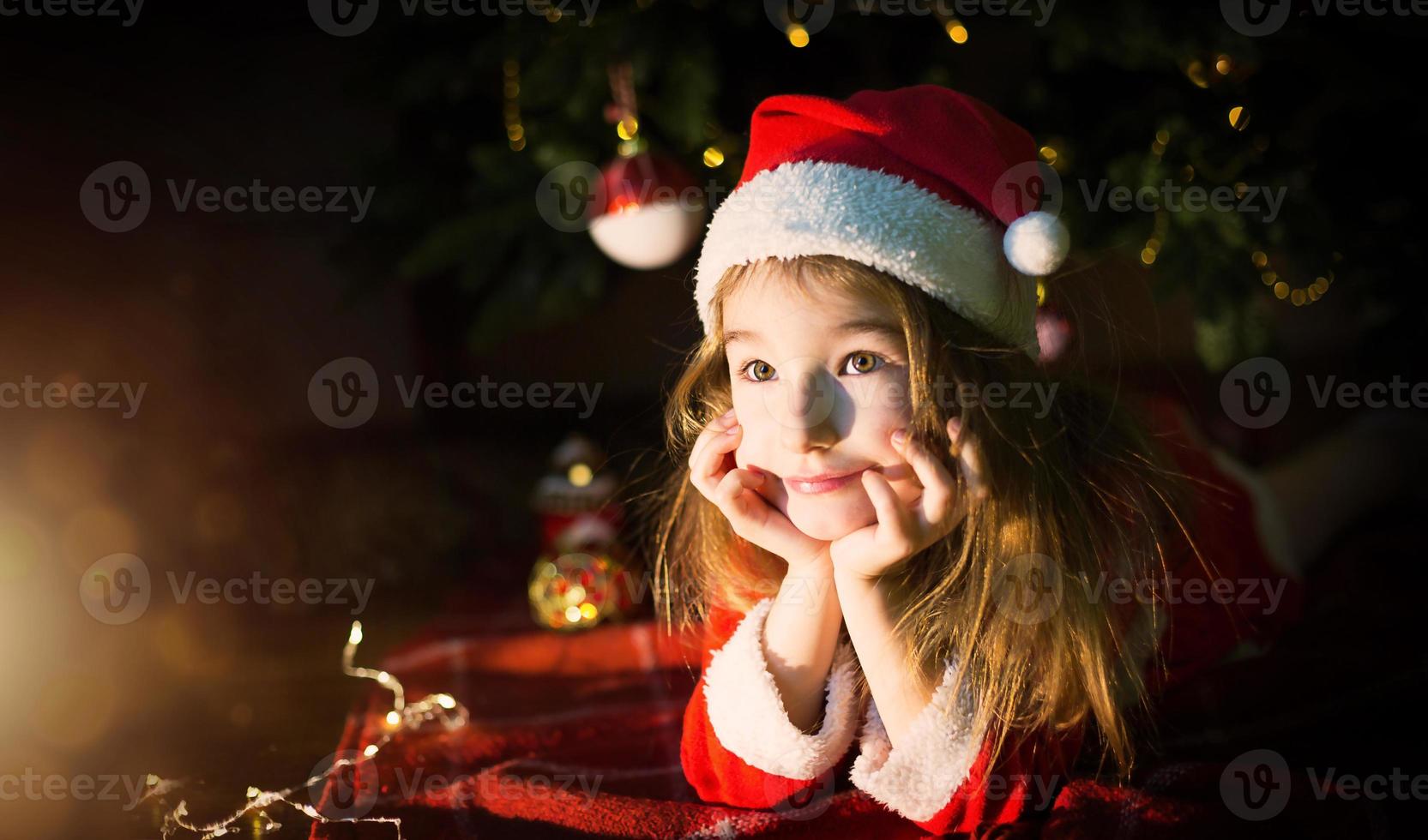 menina com um chapéu de Papai Noel e vestido vermelho debaixo da árvore de natal está sonhando, esperando o feriado, deitado sobre um cobertor xadrez. uma carta em um pedaço de papel, presentes. ano novo, natal. luzes de desfocagem foto