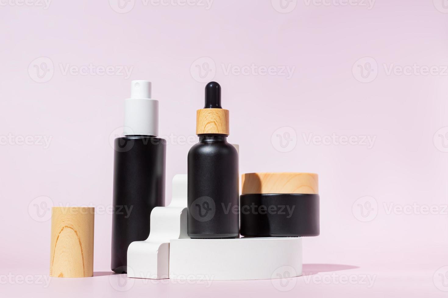 três garrafas de vidro preto com formas geométricas na moda em um fundo rosa... pedestal branco e escada. produtos para a pele, cosméticos naturais. conceito de beleza para cuidados com o rosto foto