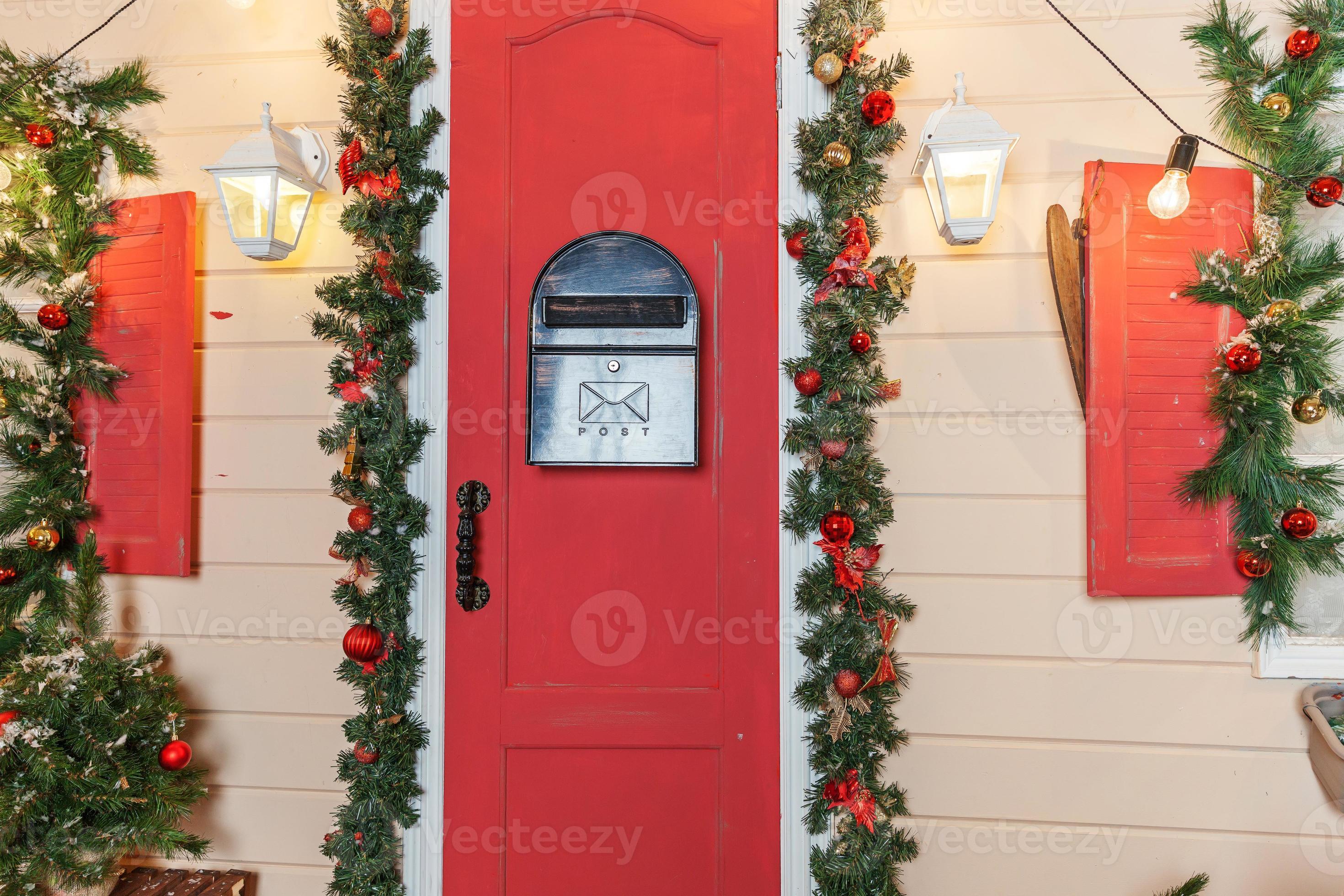ideia de decoração de varanda de natal. entrada da casa com porta vermelha  decorada para feriados. guirlanda de grinalda vermelha e verde de galhos de  árvores do abeto e luzes na grade.