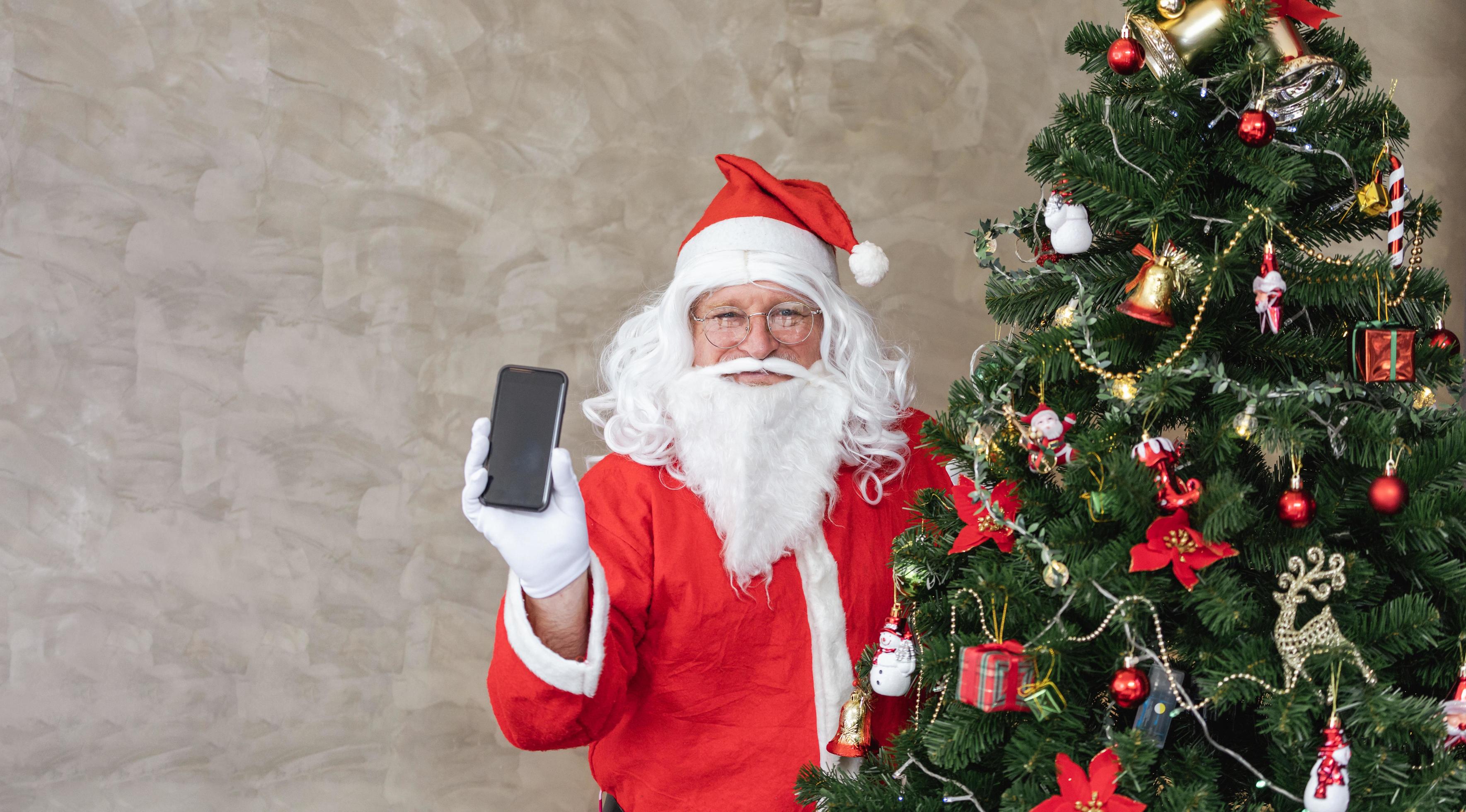 papai noel comemorando o natal com felicidade e emoção enquanto segura o  celular ao lado da árvore de natal totalmente decorada para promoção de  venda e uso de compras online 14202388 Foto