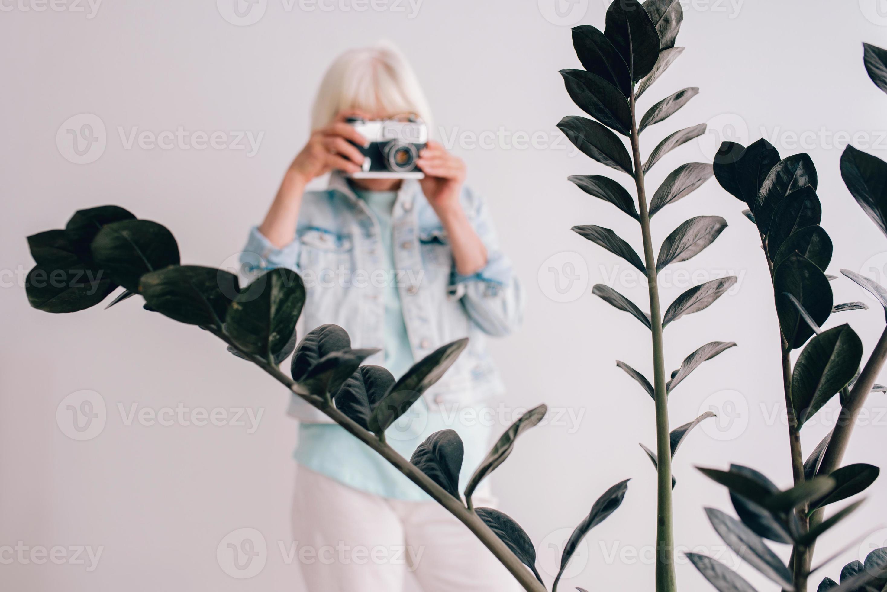 mulher elegante sênior com cabelos grisalhos e de óculos e jaqueta jeans, tirando fotos de flores com a câmera de filme. idade, hobby, anti-idade, vibrações positivas, conceito de fotografia