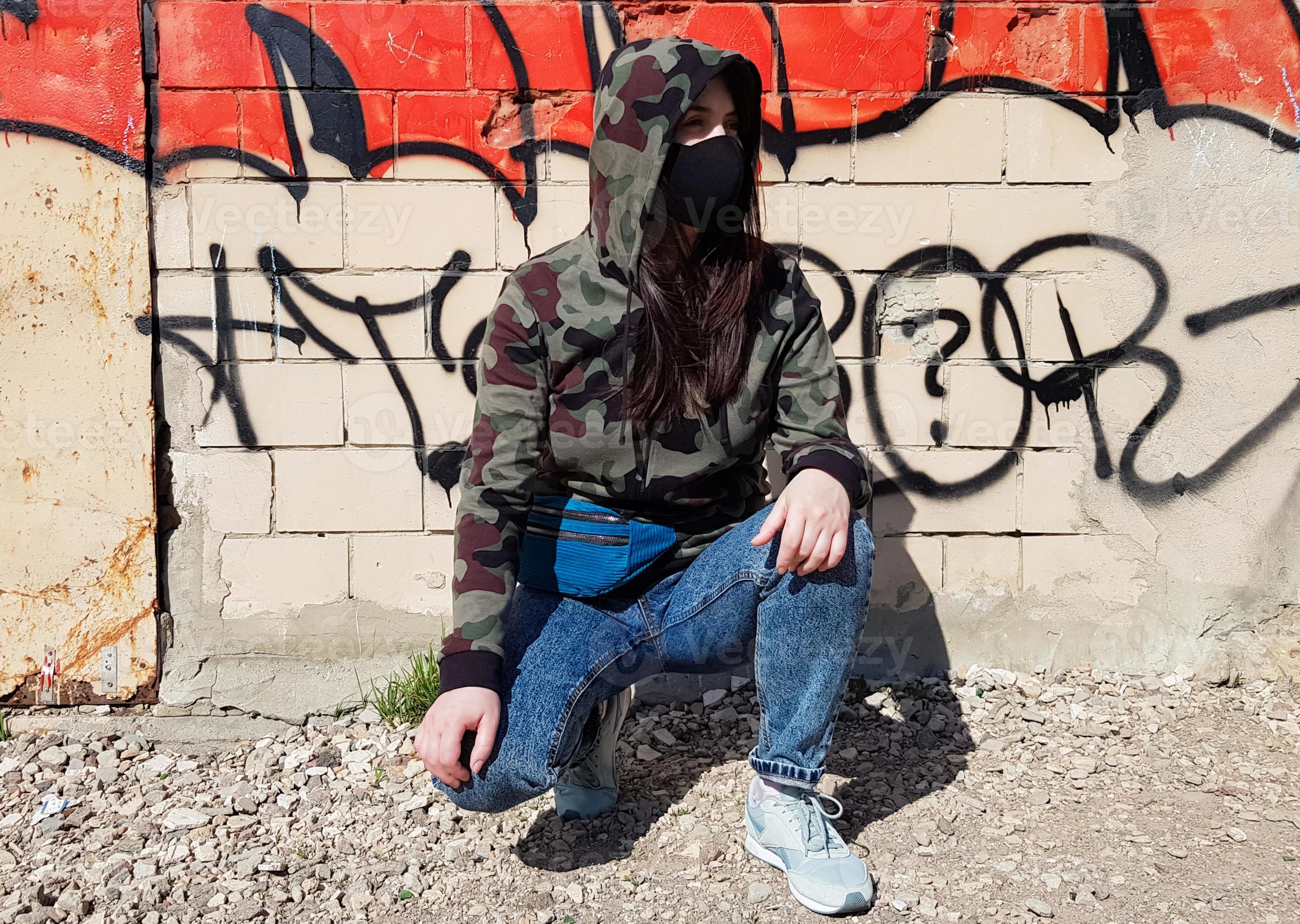 ucrânia, kiev - 24 de abril de 2020. uma bela mulher com capuz e máscara  protetora, vestida de jeans e jaqueta caqui camuflada, senta-se perto de  uma parede com pichações. na rua 4536264 Foto de stock no Vecteezy