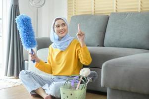 jovem mulher muçulmana feliz usando luvas amarelas e segurando uma cesta de material de limpeza na sala de estar. foto